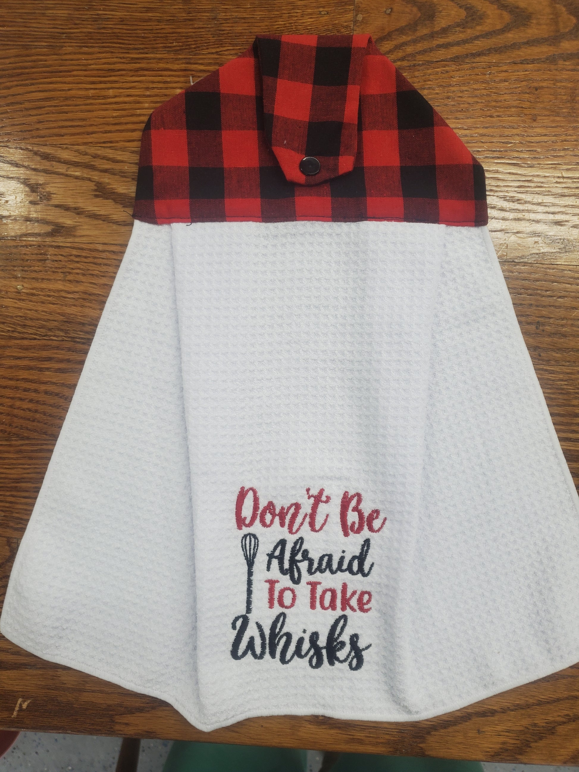 Attachable white kitchen towel with red and black plaid border and text Don't be afraid to take whisks